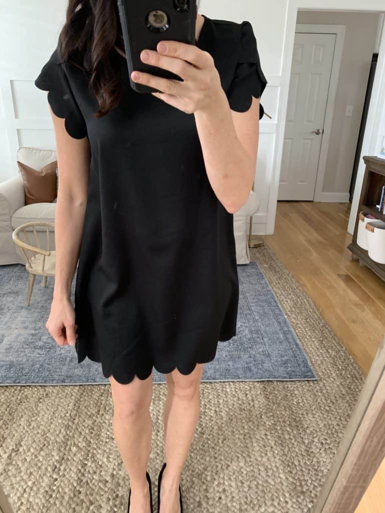 A mirror selfie of a woman wearing a black dress with scalloped cap sleeves.