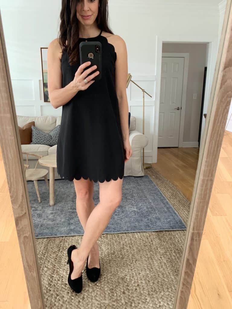 A mirror selfie of a woman wearing a black halter tie dress with scalloped hem and black kitten heels.