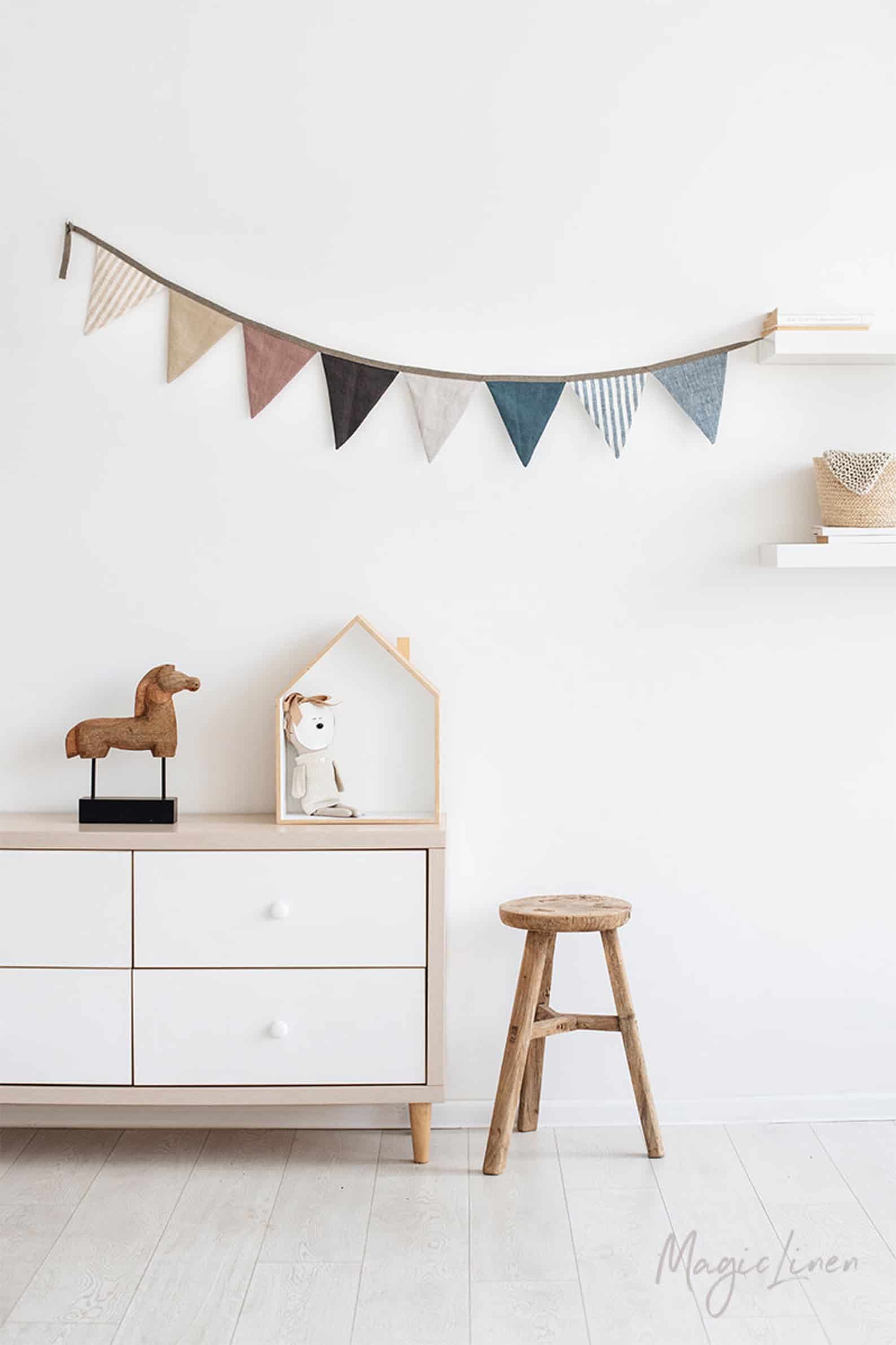 A banner of different patterned fabric flags hangs on a white wall, above a white storage cabinet decorated with a doll house and toy horse.