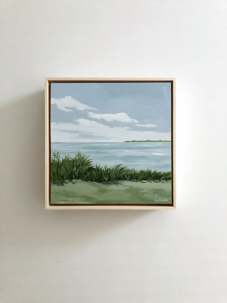 This coastal landscape artwork shows a serene green grassy bluff overlooking the ocean with white clouds in the sky. This painting is the perfect accessory in a serene and simple bedroom design.