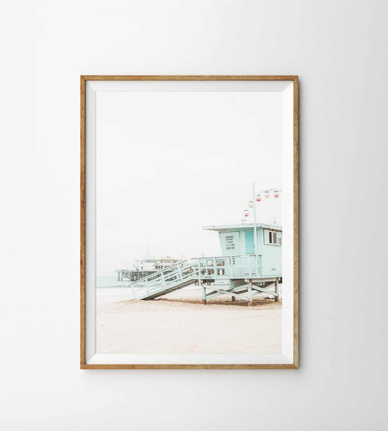 This coastal art print features a vintage lifeguard stand on an empty beach near the coast and is framed with a natural wood frame, hanging on a white wall.