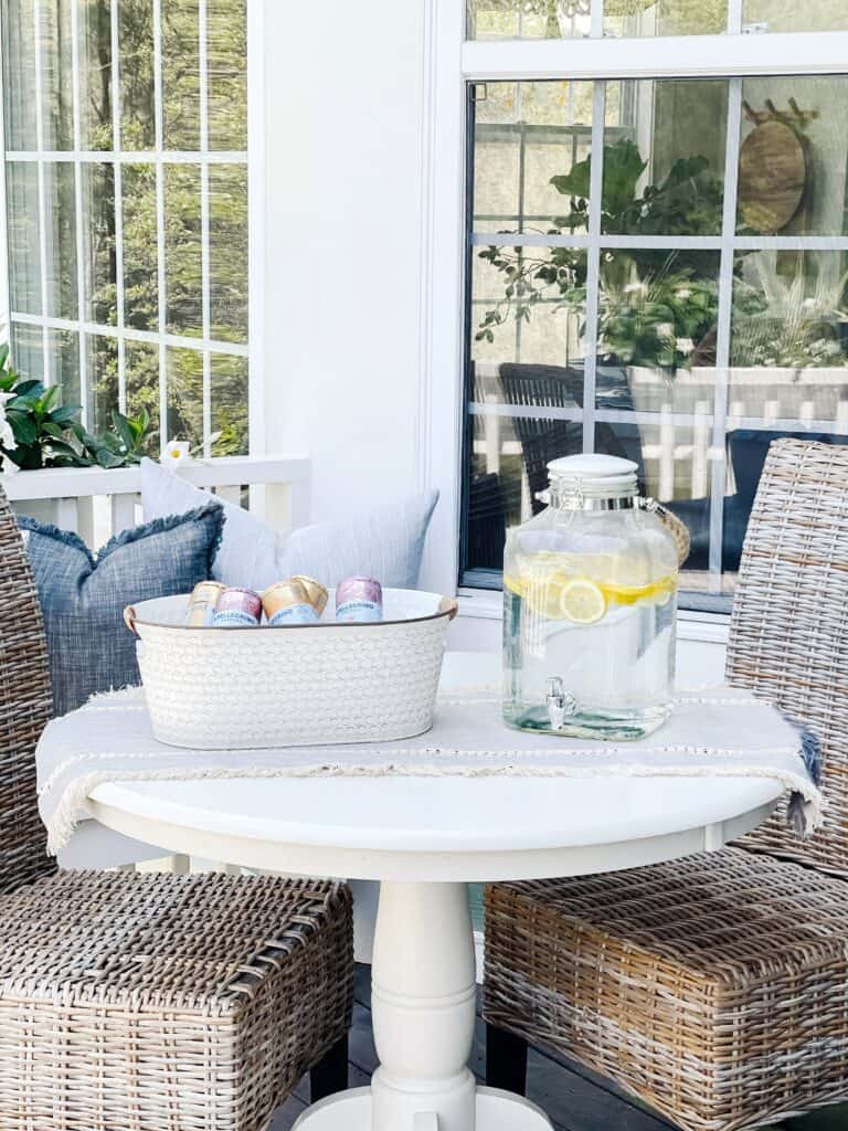Spring patio decor from Walmart. A bucket with ice and drinks, and a glass jug filled with lemon water sits on an outdoor patio table with a white table runner and woven patio chairs.