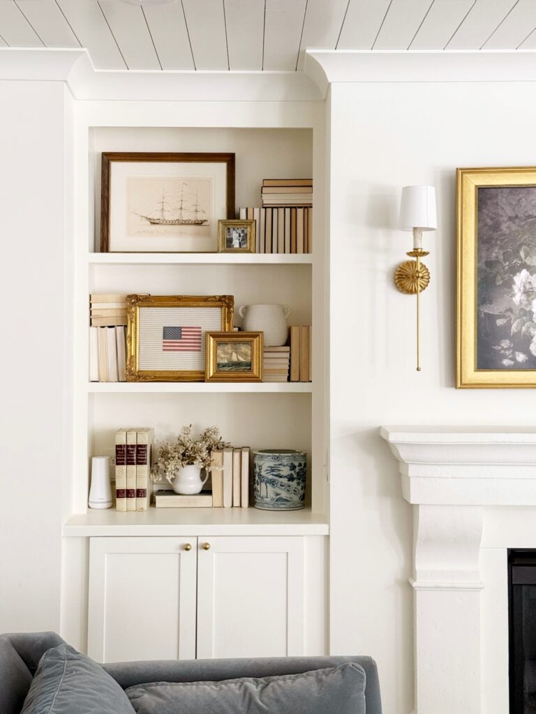 Styled built-in bookshelf with neutral-toned books, vintage art, brass picture frames, and coastal-inspired decor in a cozy living room with white shiplap ceiling and fireplace mantel.